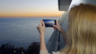 Çekici bir genç kadın, bir spa otel balkon bir smartphone kullanarak akıllı cep telefonundan bir uygulama kullanarak deniz kenarında bir gün batımı bir kadın fotoğrafını. 4k