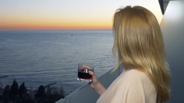 女性は、リゾートの海の景色とバルコニーに立っています。夕方には夕日を見てカクテルを飲みます。4 k — ストック動画