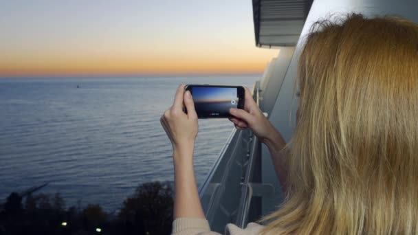 Jeune femme séduisante utilisant un smartphone au balcon d'un hôtel spa, Une femme photographie un coucher de soleil au bord de la mer à l'aide d'une application sur un téléphone portable intelligent. 4k — Video