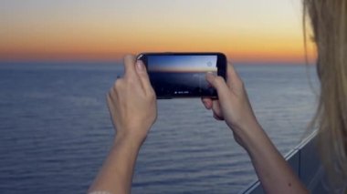 Çekici bir genç kadın, bir spa otel balkon bir smartphone kullanarak akıllı cep telefonundan bir uygulama kullanarak deniz kenarında bir gün batımı bir kadın fotoğrafını. 4 k yakın çekim