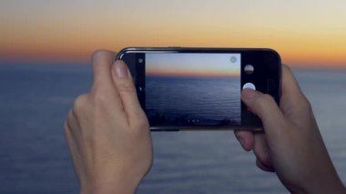 Çekici bir genç kadın, bir spa otel balkon bir smartphone kullanarak akıllı cep telefonundan bir uygulama kullanarak deniz kenarında bir gün batımı bir kadın fotoğrafını. 4 k yakın çekim