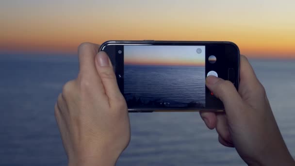 Una atractiva mujer joven usando un teléfono inteligente en el balcón de un hotel spa, Una mujer está fotografiando una puesta de sol junto al mar usando una aplicación en un teléfono celular inteligente. 4k primer plano — Vídeo de stock