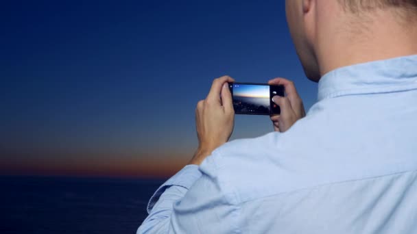 Um jovem atraente usando um smartphone na varanda de um hotel spa, um homem tira fotos do pôr do sol à beira-mar, usando um aplicativo em um smartphone. 4k close-up — Vídeo de Stock