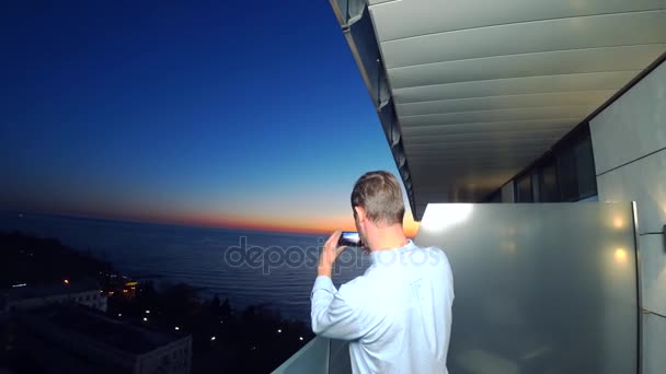 Um jovem atraente usando um smartphone na varanda de um hotel spa, um homem tira fotos do pôr do sol à beira-mar, usando um aplicativo em um smartphone. 4k close-up — Vídeo de Stock