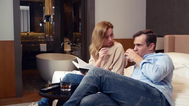 Ένα ζευγάρι άνδρα και γυναίκας καθίσει σε ένα κρεβάτι σε ένα δωμάτιο ξενοδοχείου και Συζητήστε στιγμές εργασίας, καθιστώντας τις σημειώσεις σε ένα σημειωματάριο. 4k. — Αρχείο Βίντεο