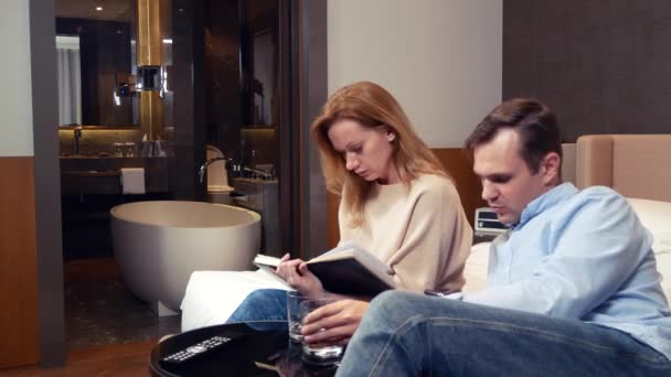 Egy pár férfi és a nő ül egy ágy, egy szállodai szobában, és megvitassák a munka pillanatok, jegyzetelés egy füzetbe. 4k. — Stock videók