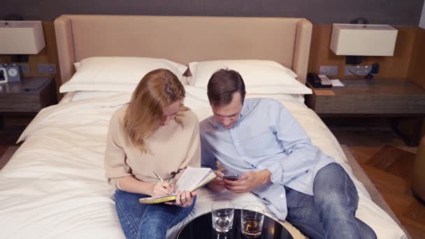 Ein paar männer und frauen sitzen auf einem bett in einem hotelzimmer, besprechen arbeitsmomente und notieren in einem notizbuch. 4k. — Stockvideo