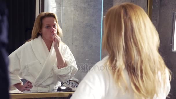 白いバスローブを着ている女性は 4 k、鏡の前で歯をブラシします。 — ストック動画