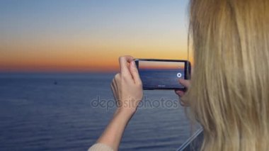 Çekici bir genç kadın, bir spa otel balkon bir smartphone kullanarak akıllı cep telefonundan bir uygulama kullanarak deniz kenarında bir gün batımı bir kadın fotoğrafını. 4k