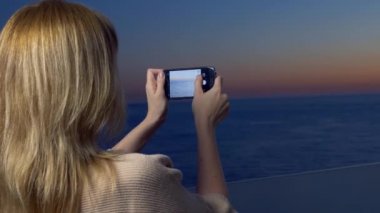 Çekici bir genç kadın, bir spa otel balkon bir smartphone kullanarak akıllı cep telefonundan bir uygulama kullanarak deniz kenarında bir gün batımı bir kadın fotoğrafını. 4k
