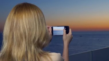 Çekici bir genç kadın, bir spa otel balkon bir smartphone kullanarak akıllı cep telefonundan bir uygulama kullanarak deniz kenarında bir gün batımı bir kadın fotoğrafını. 4k