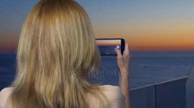 Çekici bir genç kadın, bir spa otel balkon bir smartphone kullanarak akıllı cep telefonundan bir uygulama kullanarak deniz kenarında bir gün batımı bir kadın fotoğrafını. 4k