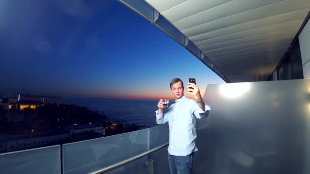 Un joven atractivo usando un teléfono inteligente en el balcón de un hotel spa, un hombre toma fotos de la puesta de sol junto al mar, utilizando una aplicación en un teléfono inteligente. 4k primer plano — Vídeos de Stock