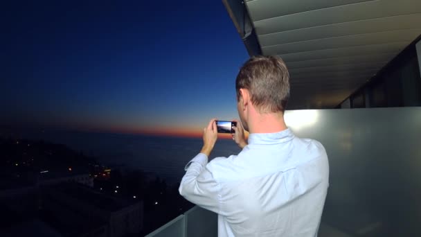 Jeune homme séduisant utilisant un smartphone sur le balcon d'un hôtel spa, un homme prend des photos du coucher de soleil au bord de la mer, à l'aide d'une application sur un smartphone. Gros plan 4k — Video