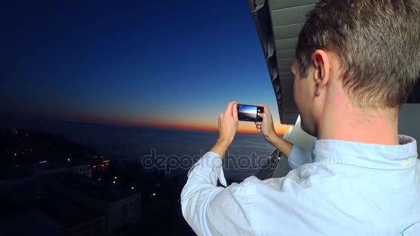 Un joven atractivo usando un teléfono inteligente en el balcón de un hotel spa, un hombre toma fotos de la puesta de sol junto al mar, utilizando una aplicación en un teléfono inteligente. 4k primer plano — Vídeo de stock