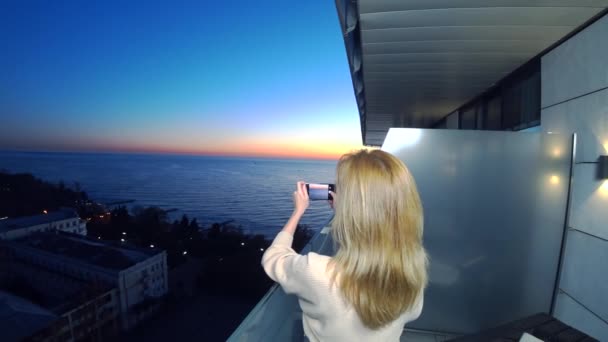 Uma jovem atraente usando um smartphone na varanda de um hotel de spa, Uma mulher está fotografando um pôr do sol junto ao mar usando uma aplicação em um telefone celular inteligente. 4k — Vídeo de Stock