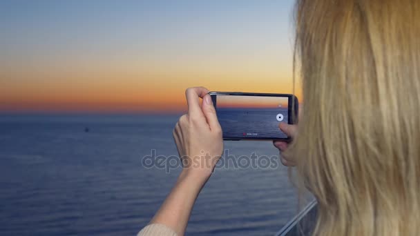 Uma jovem atraente usando um smartphone na varanda de um hotel de spa, Uma mulher está fotografando um pôr do sol junto ao mar usando uma aplicação em um telefone celular inteligente. 4k — Vídeo de Stock