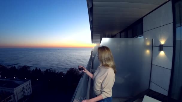 Женщина стоит на балконе курорта с видом на море. пьет коктейль вечером, глядя на закат. 4k — стоковое видео