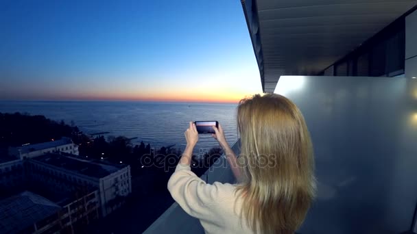 Çekici bir genç kadın, bir spa otel balkon bir smartphone kullanarak akıllı cep telefonundan bir uygulama kullanarak deniz kenarında bir gün batımı bir kadın fotoğrafını. 4k — Stok video