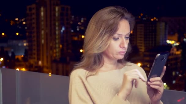 Ung, glad kvinna med en smartphone på balkongen i staden på natten, 4k — Stockvideo