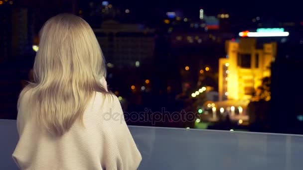 Een blonde vrouw op het terras van een resorthotel met uitzicht op de lichtjes van de stad van een nacht, een warme drank drinken uit een beker. achtergrond vervagen, 4k. — Stockvideo