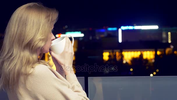 Una mujer rubia en la terraza de un hotel resort con vistas a las luces de una ciudad nocturna, bebiendo una bebida caliente de una taza. desenfoque de fondo, 4k . — Vídeos de Stock