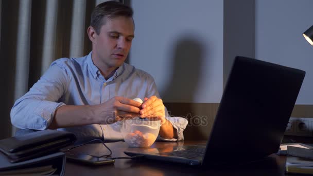 En man äter kokt kyckling ägg från en matbehållare, arbetar på en bärbar dator, sent på natten, hälsosam mat, en proteinrik kost. 4k — Stockvideo