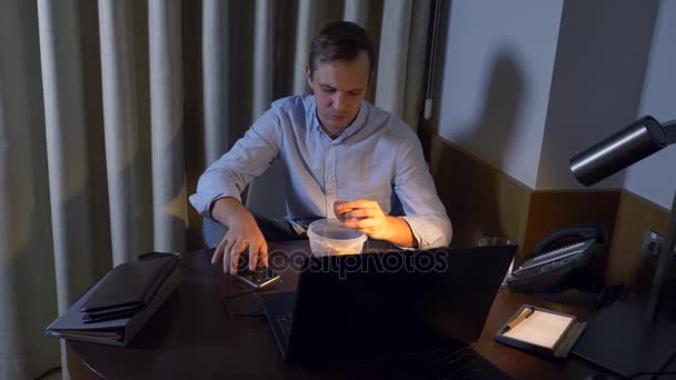 Un hombre come huevos de pollo hervidos de un recipiente de comida, trabajando en una computadora portátil, tarde en la noche, comida saludable, una dieta de proteínas. 4k — Vídeo de stock