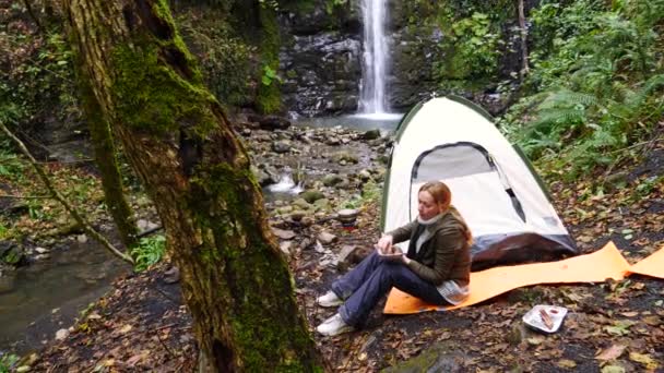 4k, slowmotion. turist flicka äter korv grill i skogen nära ett tält mot vattenfall bakgrund. — Stockvideo