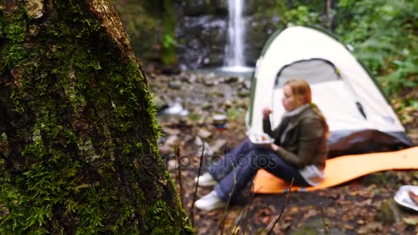 4k, cámara lenta. chica turística comiendo una parrilla de salchicha en el bosque cerca de una tienda de campaña contra un fondo de cascada . — Vídeos de Stock