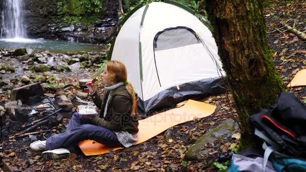 4k, Zeitlupe. Touristenmädchen beim Wurstgrillen im Wald in der Nähe eines Zeltes vor einem Wasserfall-Hintergrund. — Stockvideo