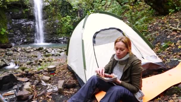 4K, câmara lenta. menina turista comer uma grade de salsicha na floresta perto de uma barraca contra um fundo cachoeira . — Vídeo de Stock