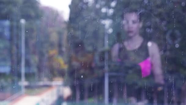 Reflet dans la fenêtre panoramique de la salle de sport avec vue sur la nature. la jeune fille court sur le tapis roulant dans la salle de gym, devant la fenêtre couverte et la pluie. 4k , — Video