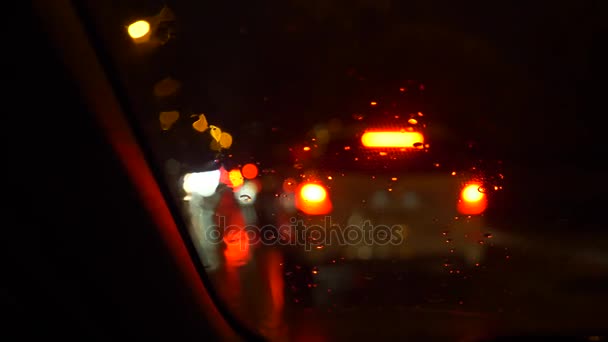 Дощові потоки та двірники на передньому скляному вікні автомобіля, що зупиняється біля дороги з красивим барвистим розмитим світлом руху на вулиці. 4k — стокове відео