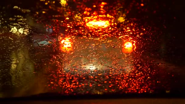 Los arroyos de lluvia en la ventana frontal de la ventana del parabrisas del coche se detienen junto a la carretera con hermosos semáforos borrosos de colores fuera de la carretera. 4k — Vídeo de stock