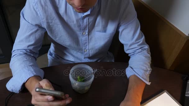 En man äter broccoli från en matbehållare, arbetar på en bärbar dator, sent på kvällen, hälsosam mat, kost. 4k. — Stockvideo