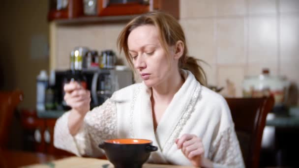 Een bleke vrouw in een badjas is het eten aan de tafel. Ze heeft een moe. 4k, achtergrond vervagen. Close-up — Stockvideo