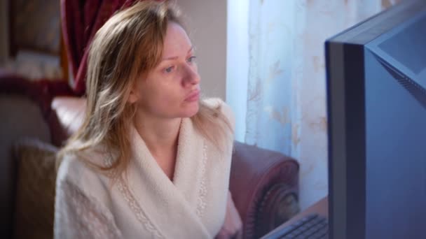 Una mujer en un albornoz y con el pelo mojado sentado en casa en el sofá, viendo los cambios en el gráfico de cambio de divisas, mirando el monitor de la computadora, freelancing, 4k, difuminando el fondo — Vídeos de Stock