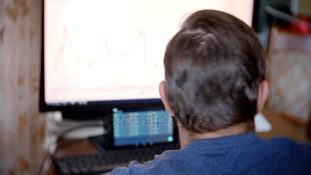 Un hombre sentado en casa en la mesa de la cocina, viendo los cambios en el gráfico de cambio de divisas, mirando el monitor de la computadora. freelancing, 4k, desenfoque de fondo — Vídeos de Stock