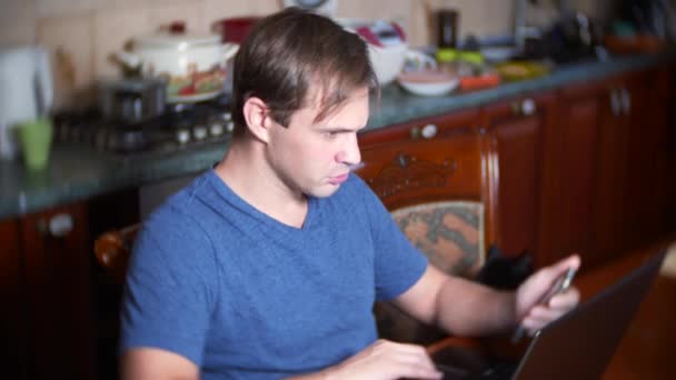 Een man zit thuis aan de keukentafel, wisselen kijken naar veranderingen in de valuta grafiek, kijken naar de computermonitor. freelancen, 4k, achtergrond vervagen — Stockvideo