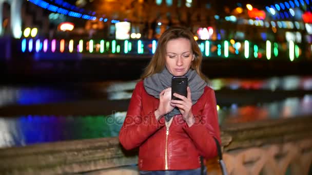 Una mujer utiliza su teléfono inteligente con una pantalla táctil. en un muelle iluminado por la noche, durante una fría noche de otoño cerca de un río, esperando a un amigo. 4k, desenfoque de fondo — Vídeo de stock