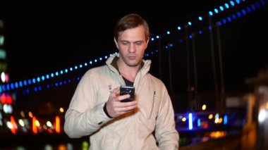 Adam onun smartphone dokunmatik ekran ile kullanır. bir gecede nehir kıyısında akşam, bekleyen soğuk sonbahar on quay, aydınlatılmış. 4k, arka plan bulanıklık