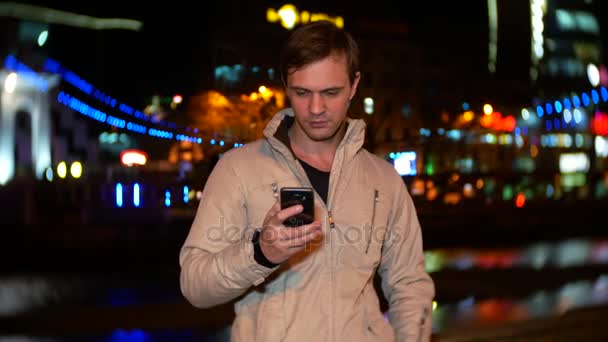 L'uomo usa il suo smartphone con un touch screen. su una banchina illuminata notte, in una fredda serata autunnale presso il fiume, in attesa. 4k, sfocatura dello sfondo — Video Stock