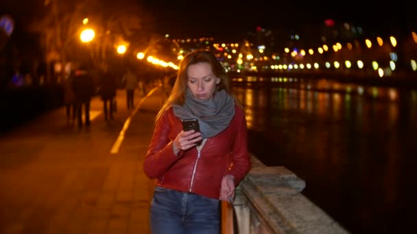 A woman uses her smartphone with a touch screen. on a night illuminated quay, during a cold autumn evening near a river, waiting for a friend. 4k, background blur — Stock Video