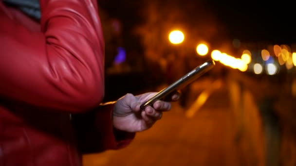 Женщина использует свой смартфон с сенсорным экраном. на ночной освещенной набережной, во время холодного осеннего вечера у реки, в ожидании друга. 4K, размытие фона — стоковое видео