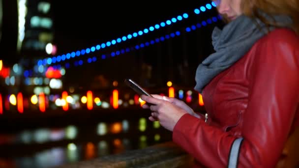 A woman uses her smartphone with a touch screen. on a night illuminated quay, during a cold autumn evening near a river, waiting for a friend. 4k, background blur — Stock Video