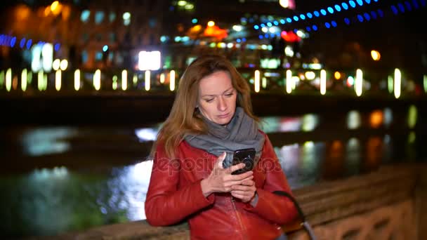 Una mujer utiliza su teléfono inteligente con una pantalla táctil. en un muelle iluminado por la noche, durante una fría noche de otoño cerca de un río, esperando a un amigo. 4k, desenfoque de fondo — Vídeos de Stock