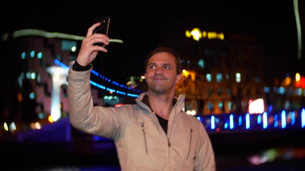 Чоловік використовує свій смартфон з сенсорним екраном. на нічній освітленій набережній, в холодний осінній вечір біля річки, чекає. 4k, розмивання тла — стокове відео