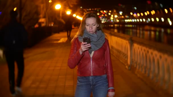 A woman uses her smartphone with a touch screen. on a night illuminated quay, during a cold autumn evening near a river, waiting for a friend. 4k, background blur — Stock Video