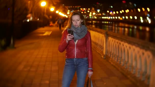 A woman uses her smartphone with a touch screen. on a night illuminated quay, during a cold autumn evening near a river, waiting for a friend. 4k, background blur — Stock Video